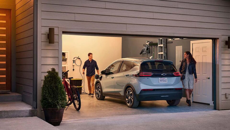 A man inside his garage has a car charger in his hand, walking toward his car while a woman walks behind the car.