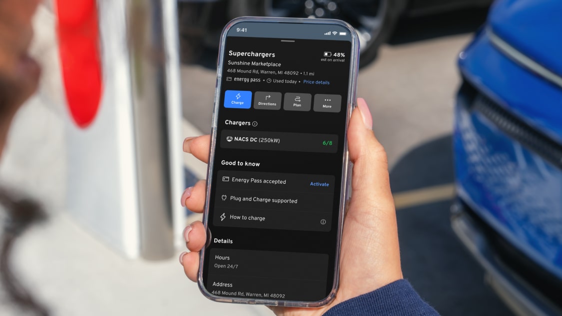 Hand holds phone by a public EV charger showing a current session dashboard in the GM vehicle mobile app with Energy Pass
