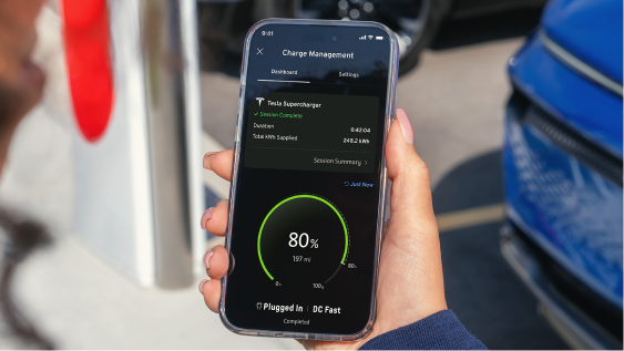 Hand holds phone by a public EV charger showing a current session dashboard in the GM vehicle mobile app with Energy Pass